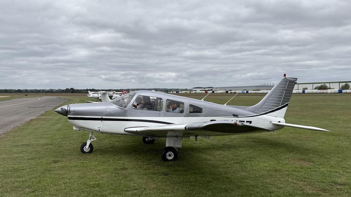 A light aircraft, which is grey and white, on the grass ready for take-off at an airfield in Leeds.