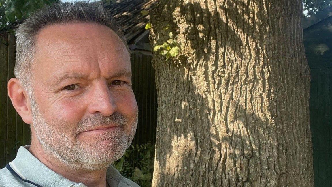 Dean Scopes stands in his garden next to one of his protected oak trees. Mr Scopes, who is smiling, has grey hair and a beard.