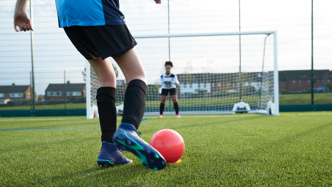 A footballer is about to take a penalty, with a foot about to strike a red football. The goalkeeper waits between the posts.