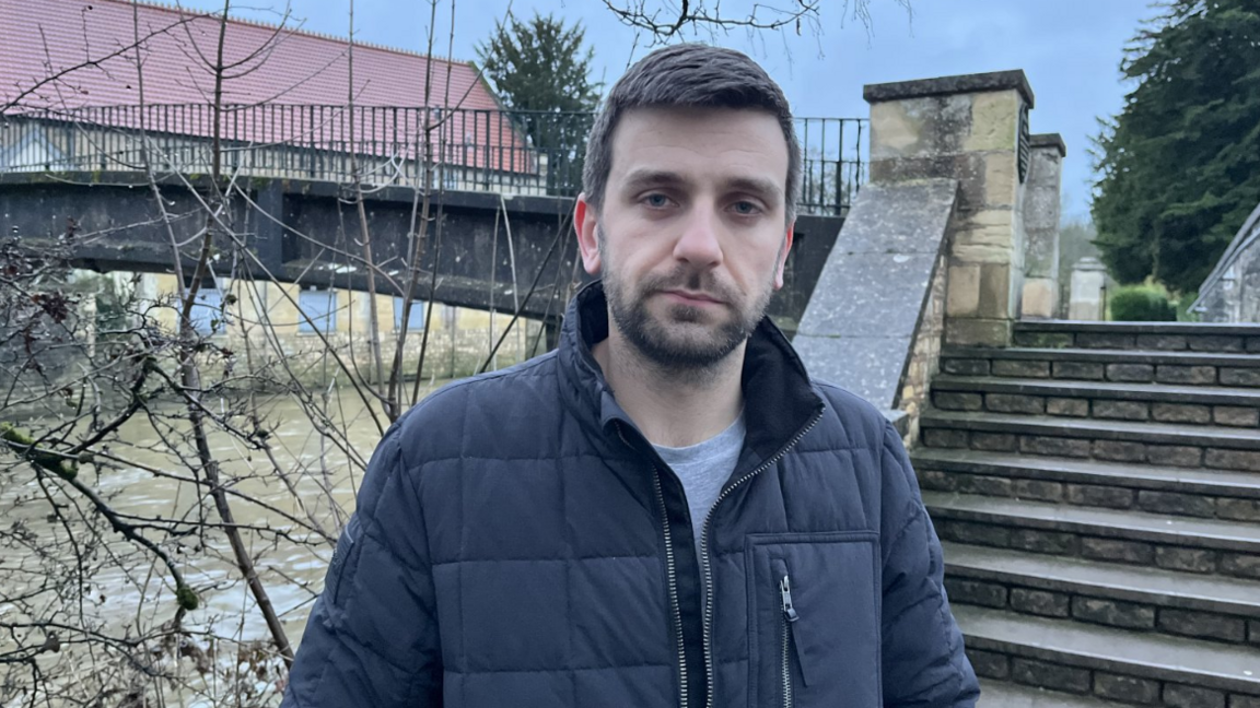 Jack Vittles looks solemnly at the camera. He is wearing a navy jacket. Behind him is a stone bridge with a brown, high river flowing beneath it. 