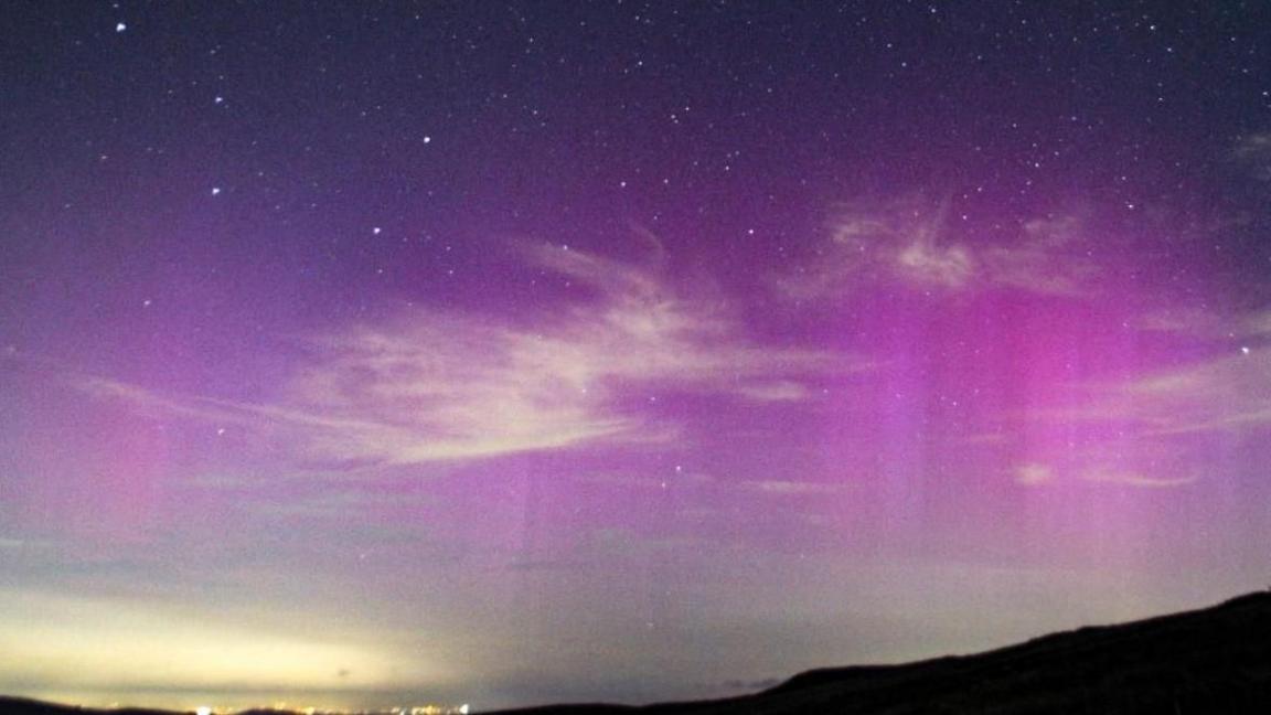 silhouette of a hill and some bright city lights at the bottom of the photo with streaks of purple and pink of the aurora in the sky.