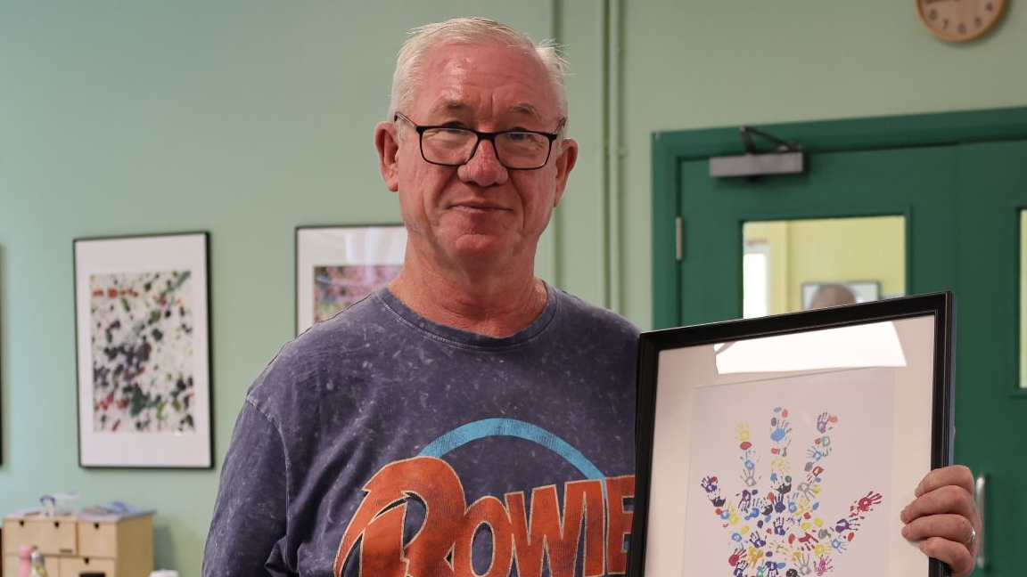 Alec is stood in front of a green wall and green door. He is smiling. He is wearing a grey David Bowie t-shirt with red writing which reads 'Bowie'. He is holding a piece of artwork which depicts a hand made up of smaller hands.