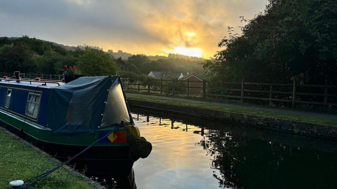 A canal boat is moored by a rope to a grassy towpath on a canal as the sun sets overhead, lighting up the sky and the canal.