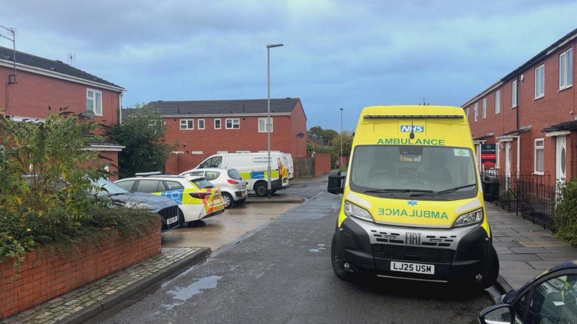 Hartington Close is made up of terraced red-bricked houses. There is a yellow ambulance parked up, along with a white "crime scene investigation" van.