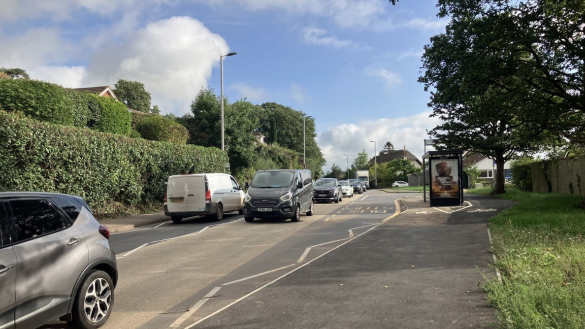Stationary traffic on the road in Pinhoe. There are cars queuing on the roads, and the pavement on the right has a bus stop on it and grass behind it. 