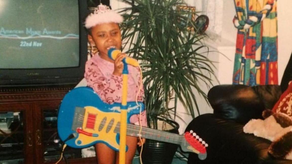 A girl is wearing a pink and red long-sleeved top and a pink tiara on her head. She is holding a yellow plastic microphone with a blue guitar at her waist. A television and plant are in the background of the living room.