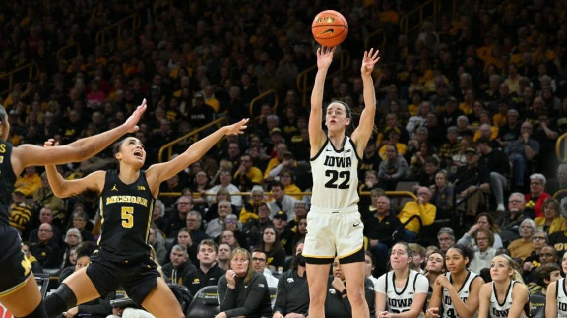 WNBA basketball player Caitlin Clark shoots a shot in a packed stadium, while other players try to block her shot. 