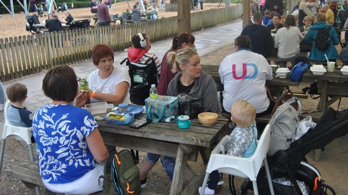 Families sitting around wooden tables at an outdoor play area.