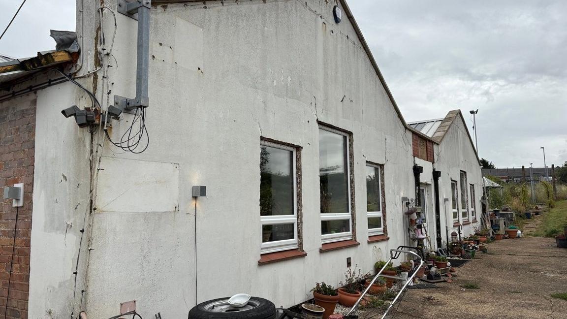 White facade of a single-storey building with square windows with white frames. There are two sections, each with a pitched roof. There are tyres and what appears to be the handle of a lawnmower on the ground in front of the building.