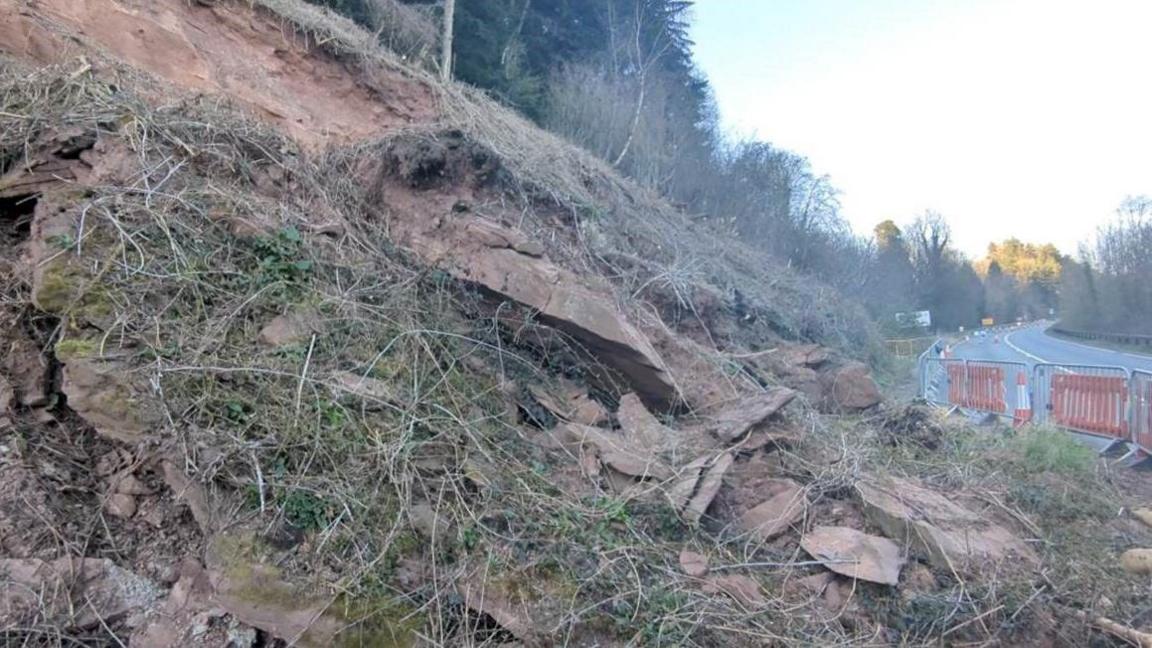 A slope of rocks near to a main road. The area has been cordoned off by gates and orange cones
