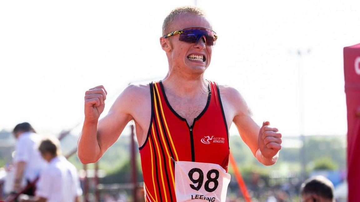 Corrin Leeming grits his teeth as he crosses a finish line. He holds both hands up by his sides in fists. He wears a red vest and wraparound sunglasses.