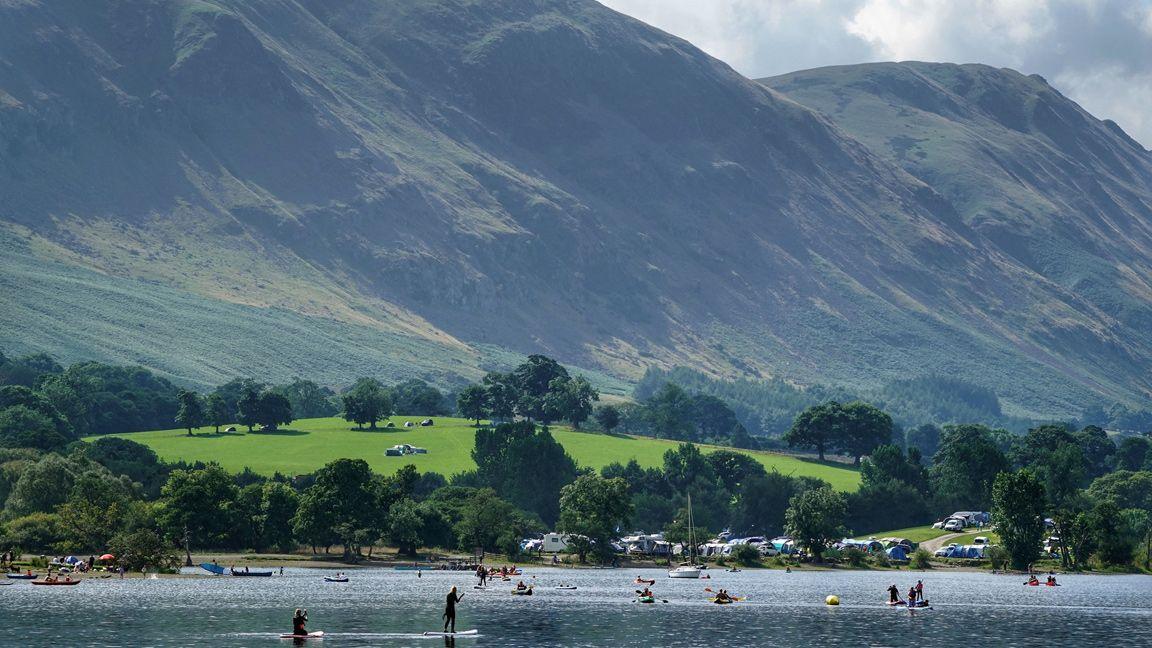 A view of Ullswater in the Lake District on a summery day. The lake is in the foreground, with people paddleboarding, kayaking and rowing. On the far bank, on a small green hill, is a campsite packed with tents and motor home. In the background, two high mountains sweep down towards the water.