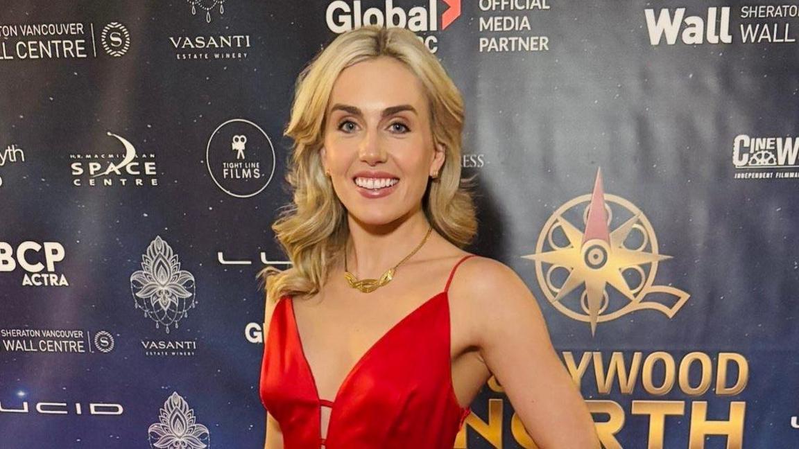 Gigi Neil, with long blond hair, smiling in a red dress, in front of a board with movie sponsor names written on it