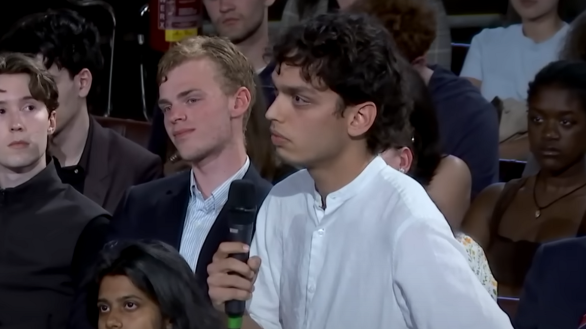 Kai Bevan standing in a white shirt at a wooden table. He has dark hair and is holding a microphone. He is looking towards Charlie Kirk who is stood opposite him.