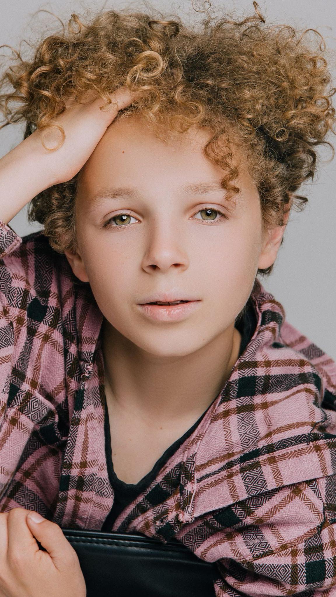 A professional-style headshot of a young boy looking into the camera while pushing a mop of curly hair away from his forehead.