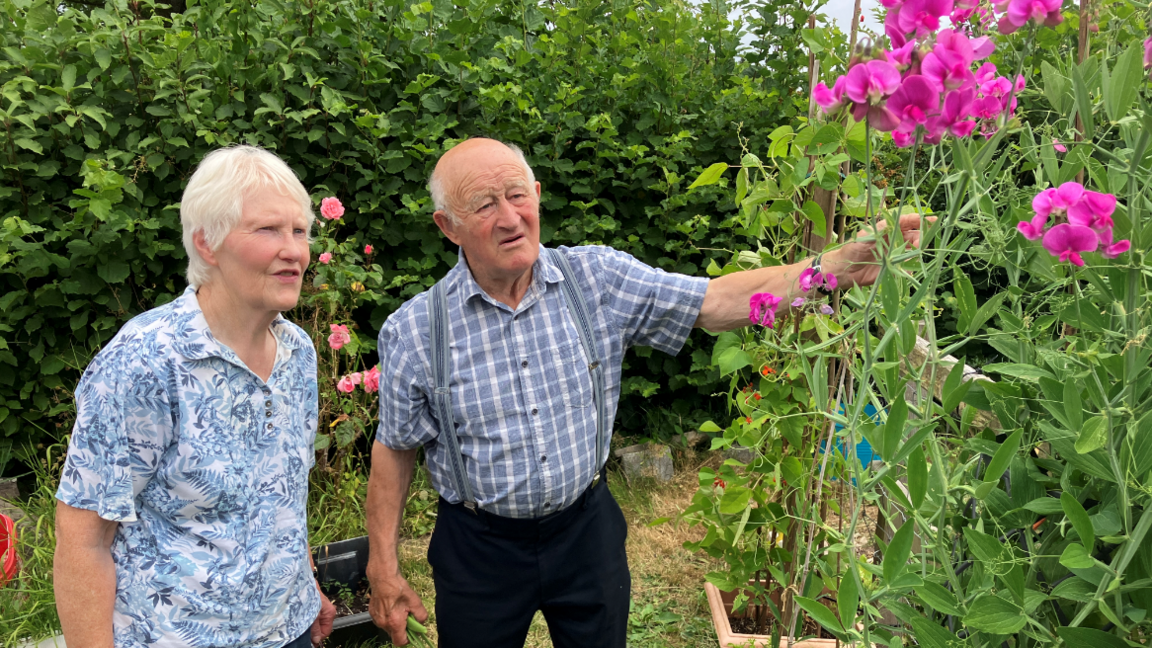 A couple stand in their garden chatting, looking at plants growing in beds.