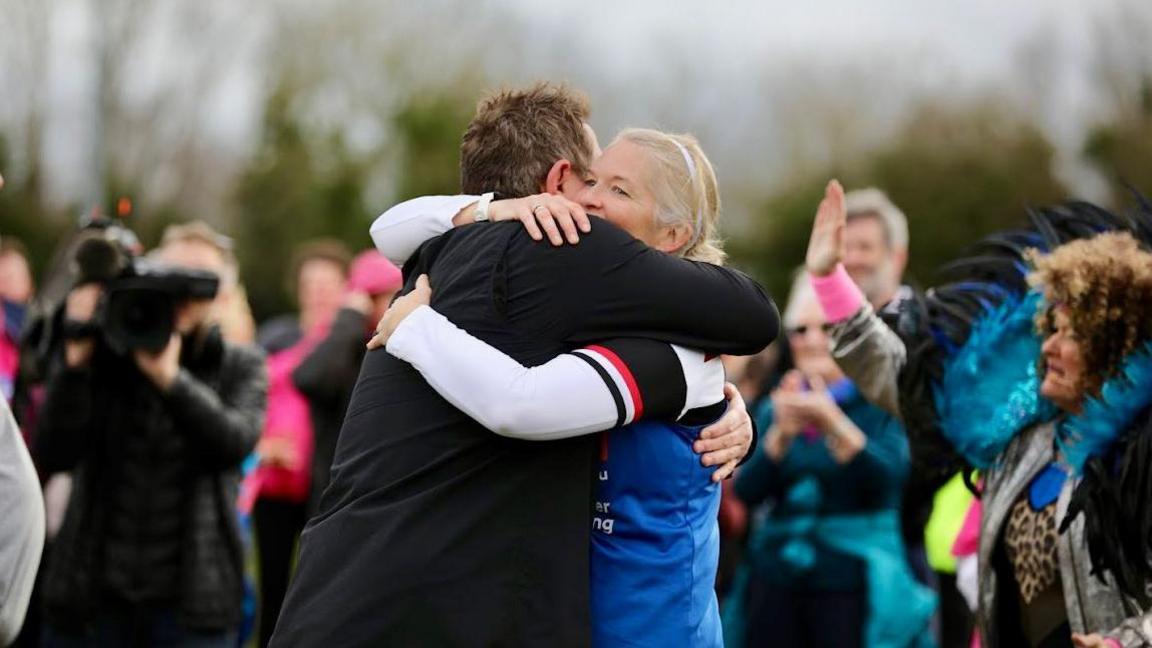 Rachel Clark hugging a man. She has blonde hair tied in a ponytail and is wearing a blue WHY charity top with a white long sleeved top underneath. Spectators can be seen in the background of the shot, with many clapping and smiling. 