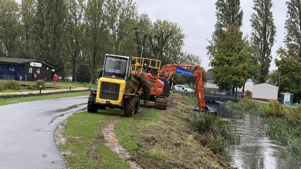 A yellow tractor behind an orange digger, which is digging in a water way. the water is banked with reeds and grass. In the distance on the right is wooden hut.