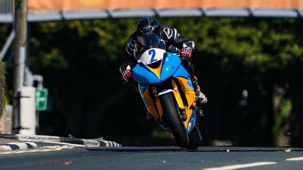 The blue and yellow motorbike of Chris Cook, which has he number two on the front in blue, leaning slightly over during the Senior race. Cook is wearing black leathers and red gloves.