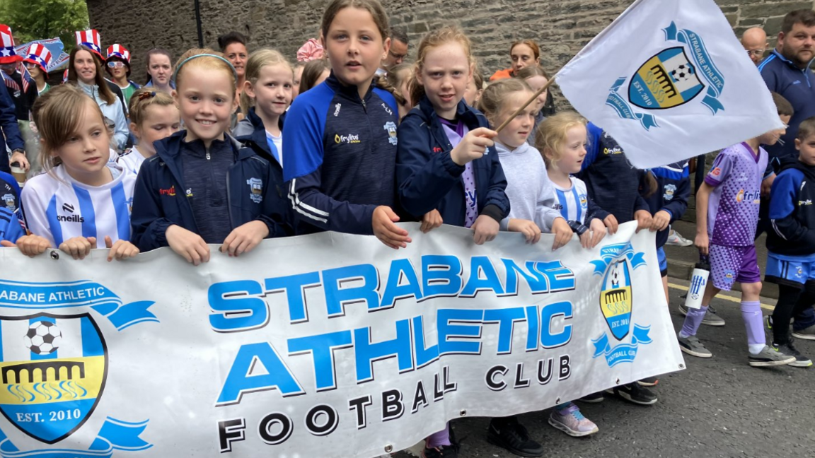 Shows girls in football tops carrying the banner of Strabane Athletic Football Club