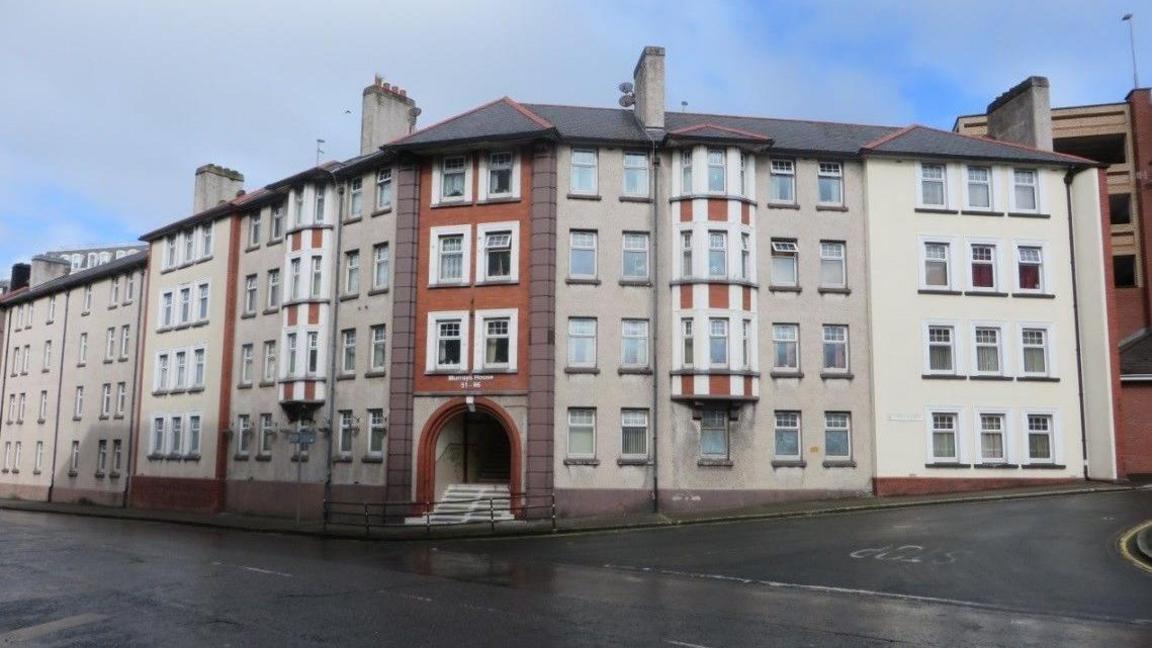 A long, four story housing block on the corner of a street junction. the entrance on the corner has a set of steps leading up to an arched entrance. 