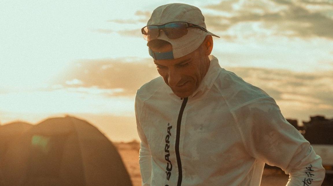 Jon Shield is wearing a cap backwards, with sunglasses resting on it. He is looking towards the ground.