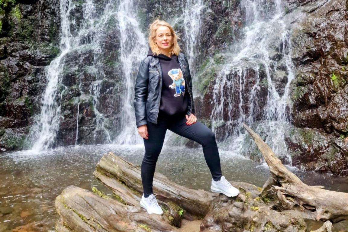 Ms Jucaite stands on an overturned tree trunk in front of a waterfall. She has long blonde hair and is wearing a black hoodie with a teddy bear picture on it, a black jacket, black jeans, and white trainers.