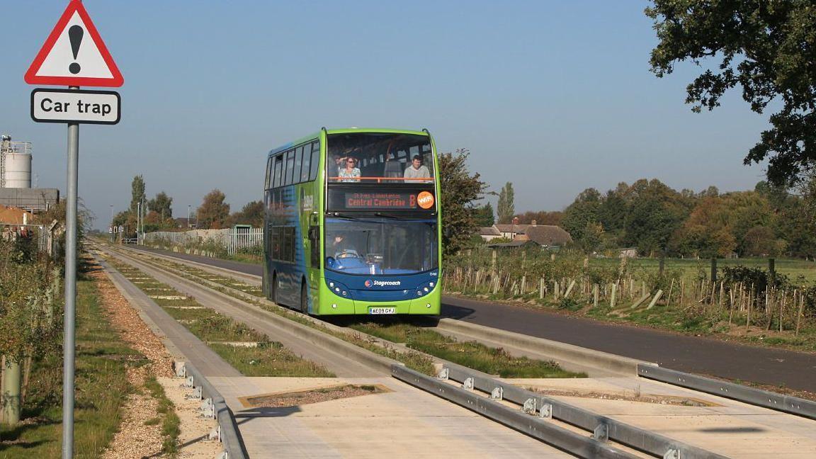 A green and blue double-decker bus heading to "Central Cambridge" on the guided busway tracks, which are like concrete railway lines that the buses fit into. A sign warning of a "car trap" - to stop cars going on to the lines - is in the foreground.