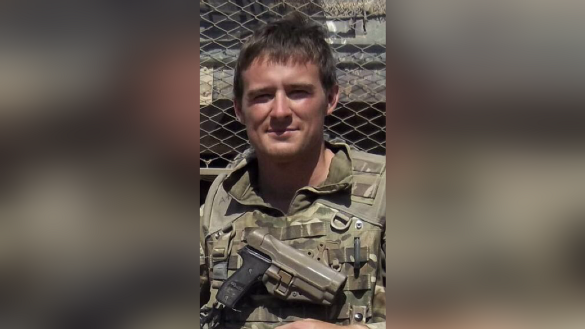 A young man in army uniform smiles with a pistol strapped across his chest