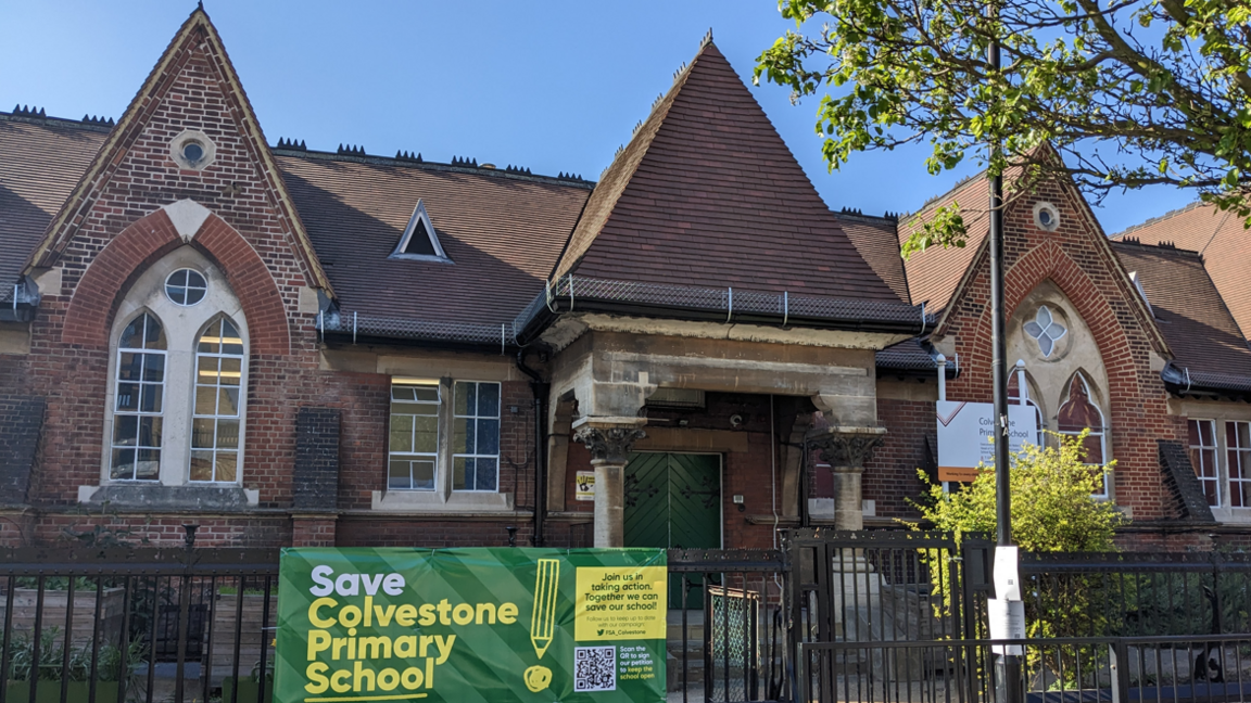 A file image of Colvestone Primary School, an old Victorian-style building with arched windows and two large green wooden doors with pillars at the entrance. In front of the building sits a black iron railing with a green and yellow tarpaulin banner reading 'save Colvestone Primary School'