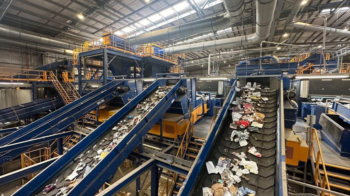 The interior of a large building with several large conveyor belts with rubbish on them and metal walkways and stairs surrounding them