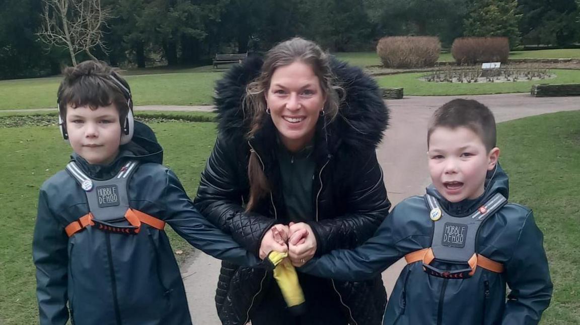 Heather Smith holds her twin boys' hands as they stand either side of her on a path in a park. One of her sons, who both have dark hair, is wearing ear protectors, and both are wearing harnesses on top of their coats. 