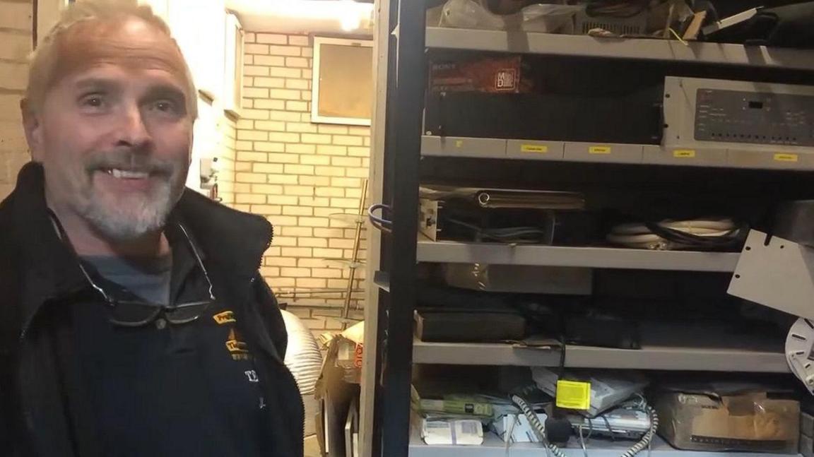 John Silk with short white hair and slight beard, wearing glasses on a string and a black fleece. There are metal shelves alongside him with various bits of equipment in storage. There are bricks and a window behind him.