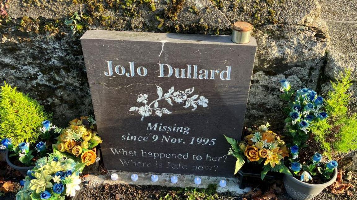 A memorial stone positioned along the side of the wall, surrounded by flower pots. It reads "JoJo Dullard Missing since 9 Nov. 1995 What happened to her? Where is JoJo now?"