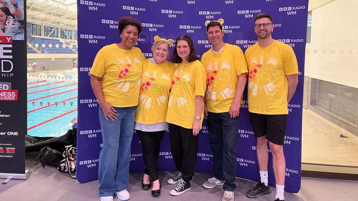 Five people wear yellow t-shirts in front of a purple board with BBC Radio WM branding. The pool is visible in the background to the left.
