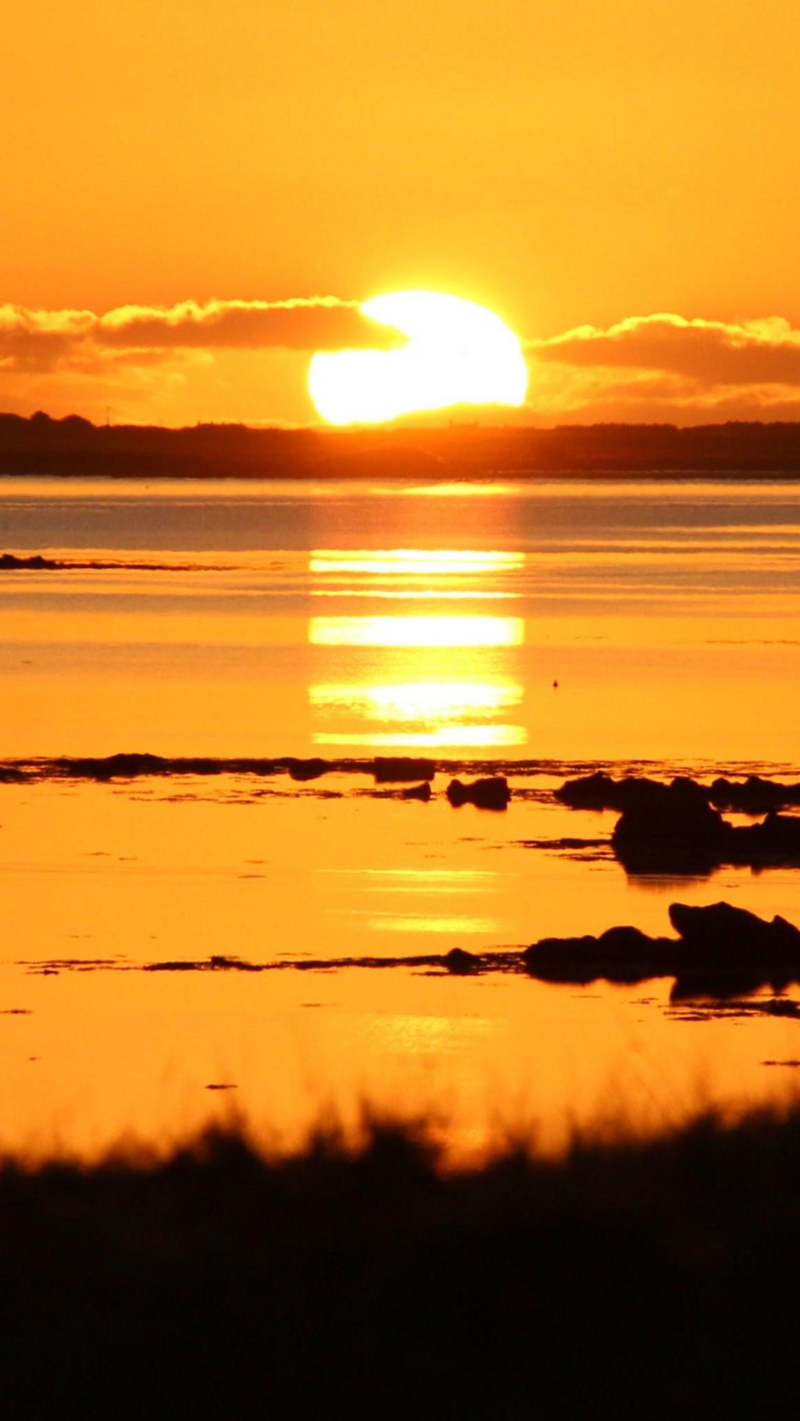 A bright orange sun reflects on calm water with scattered rocks in the foreground.