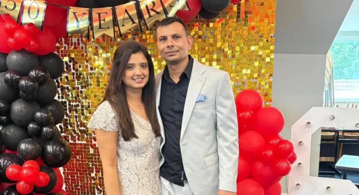 A woman in a white dress stands next to a man wearing a blue shirt and grey blazer, their hands around each other, in front of a gold backdrop.