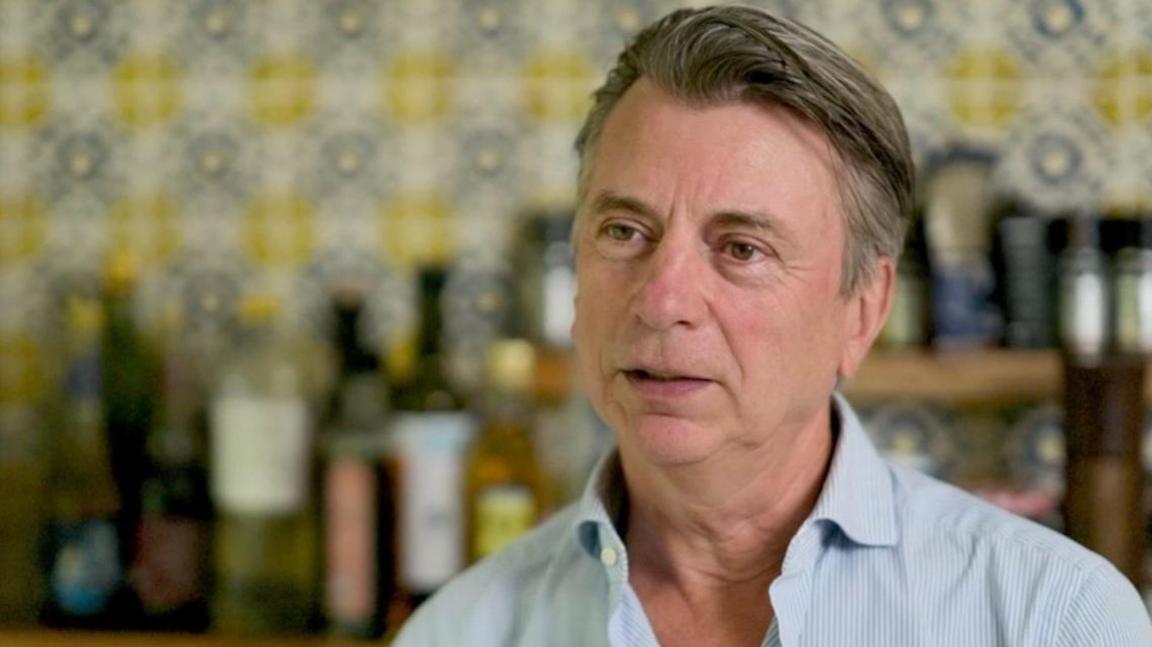 Michael Moszynski is wearing a subtly blue striped shirt in the background is an interior with different coloured bottles on a shelf and bright wallpaper.