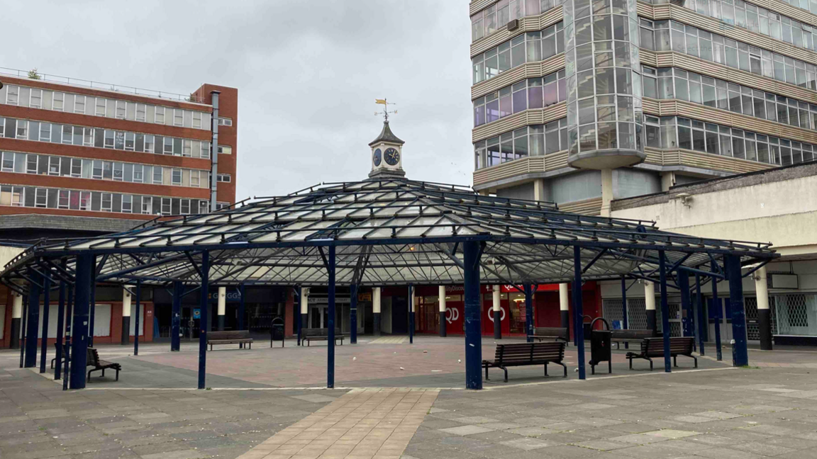 Anglia Square, a view of concrete paving, a central glass covered seating area and 1960s-style multi-storey office buildings in the background.