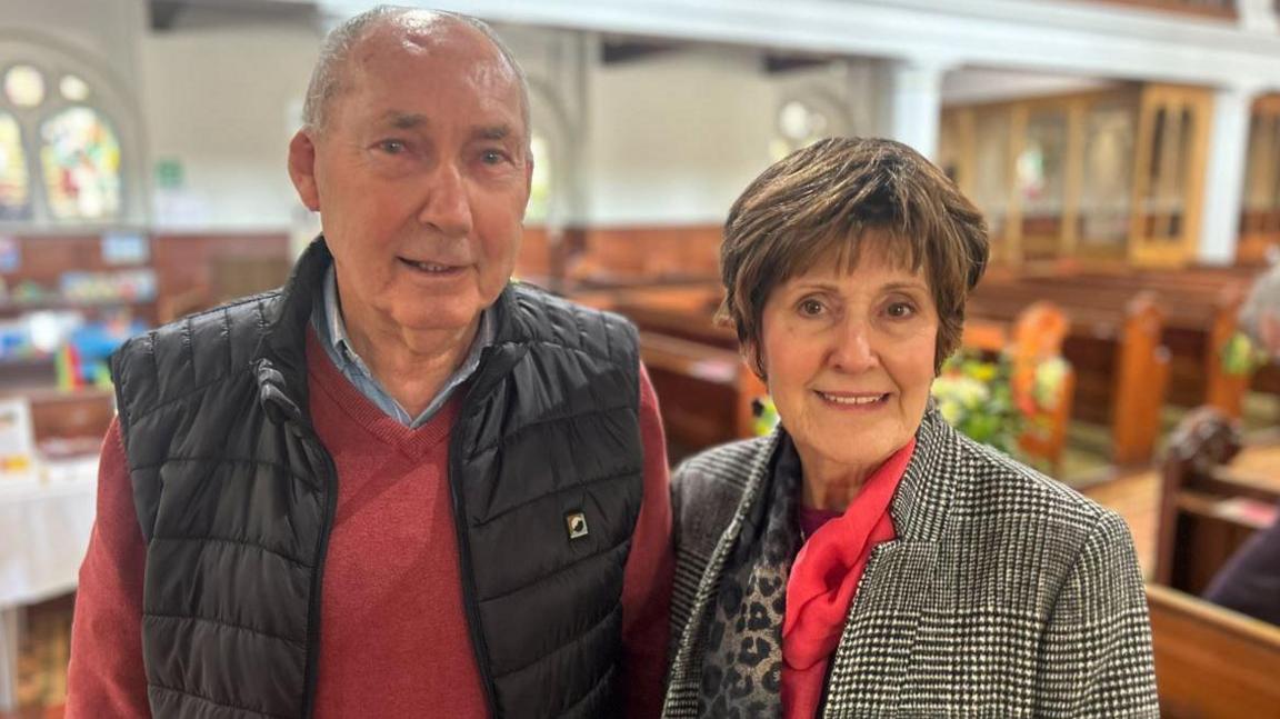 Bernard McPhee and his wife Doris smiling. They are standing in front of church pews in St George's. Doris is wearing  black and white checked coat and pink scarf, and has short brown hair. Bernard is wearing a a red jumper, blue shirt and black gillet.