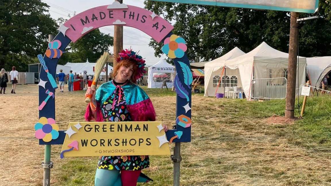 Kim Searle is standing behind a frame that says "I made this at Green Man workshops." In the background, there are white festival tents on dry grass. 