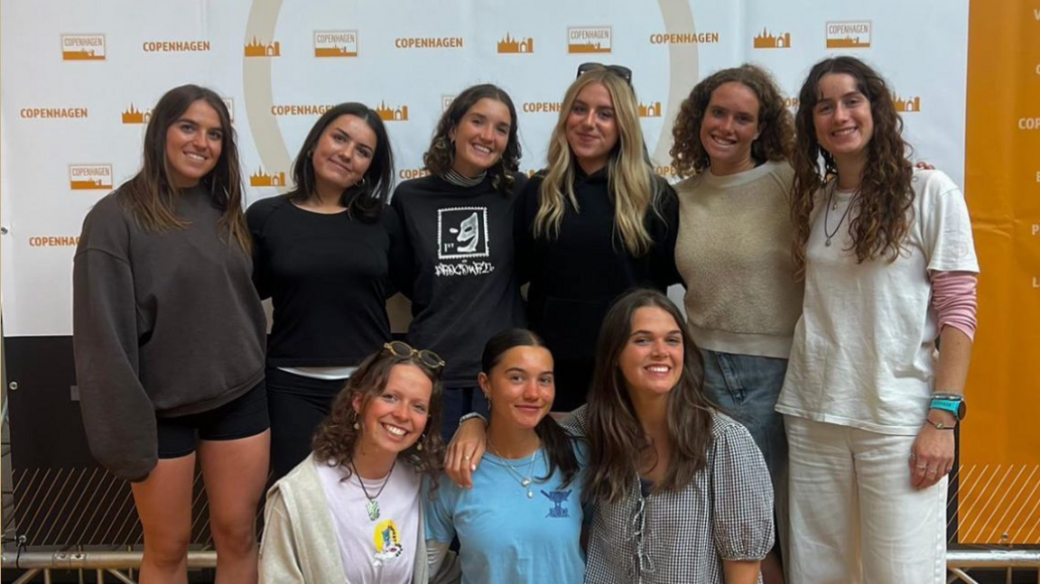 Nine 22-23 year old women smiling in front of a Copenhagen sign. 