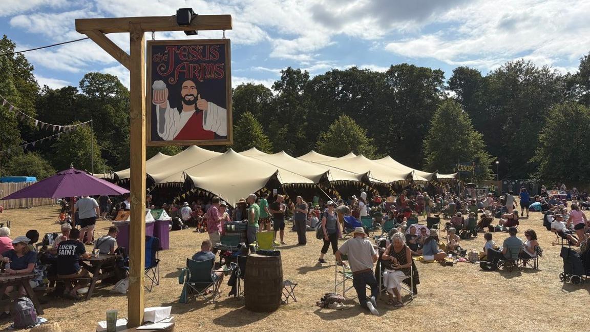 A gallows-like signpost from which a Jesus Arms sign is hanging, showing a man with dark hair and a beard holding a pint and giving a thumbs-up. There are people sitting on the grass behind and a multi-pointed marquee beyond. There are trees in the background.