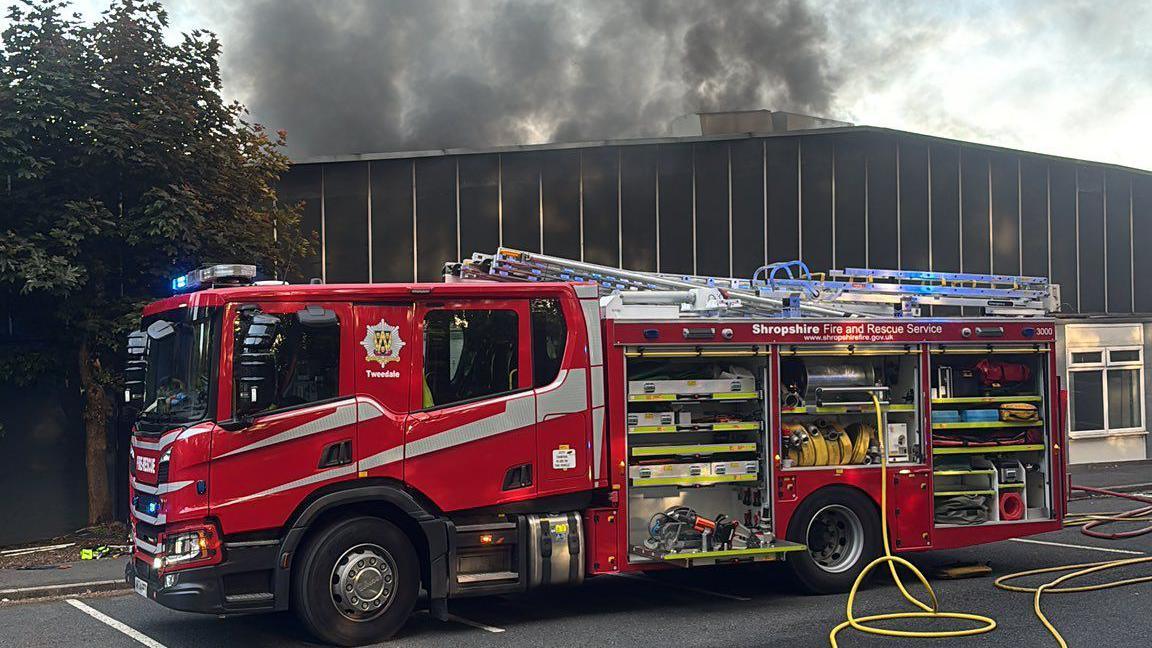 A fire engine is outside a factory warehouse building with large clouds of thick, black smoke above it. There is a yellow hose leading from the engine.