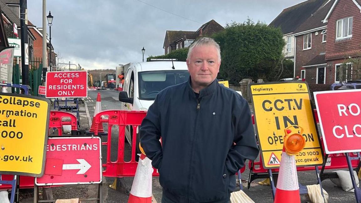 A middle-aged man with short white/grey hair. He is stood in front of a number of road closure signs in a residential street, with his hands in the pockets of his dark blue jacket. He is looking off to the right of the camera