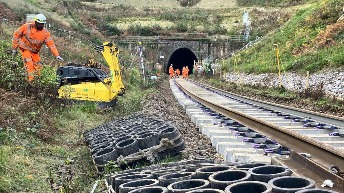 A rail track is seen leading to Honiton Tunnel, with workers both on the track and one worker operating machinery to the left. 