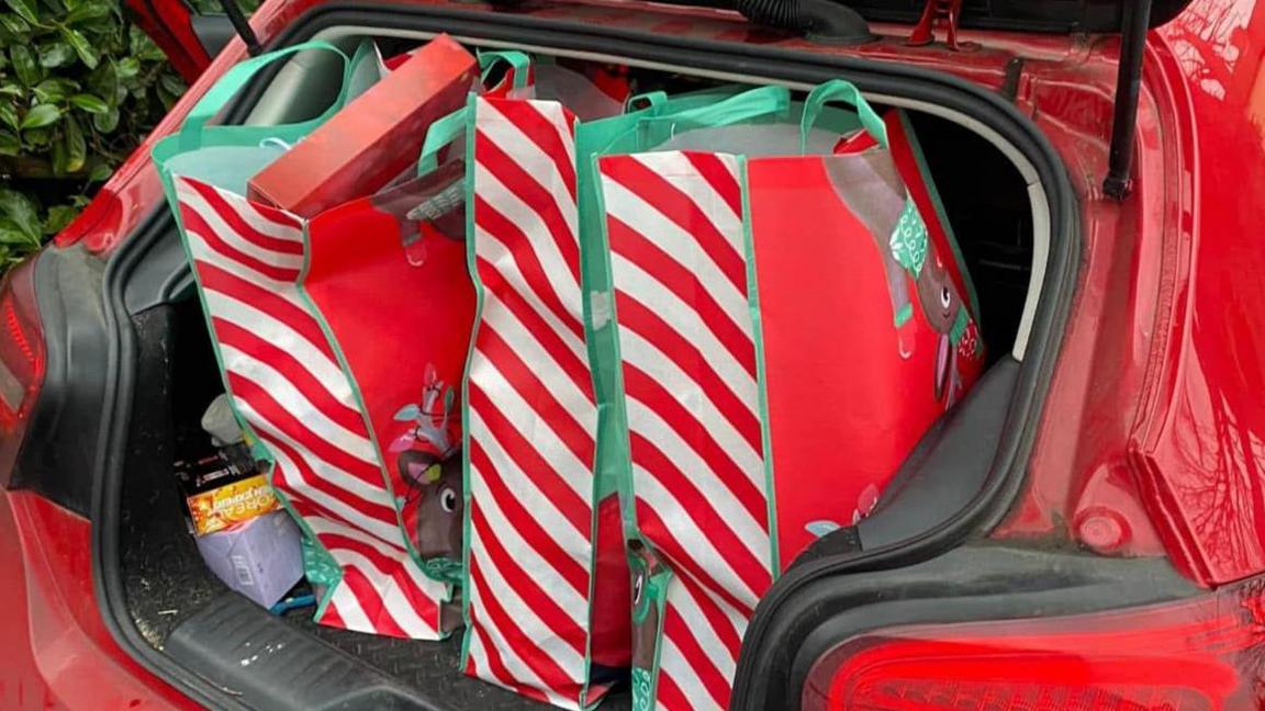 An open boot compartment of a parked red car. The boot is full of red and white striped shopping bags with reindeers on the front.