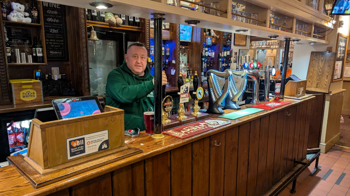 A man in a green jumper and short dark hair stood behind a bar with a number of pumps, pulling a pint. There is also a til on the bar and a number of bar towels