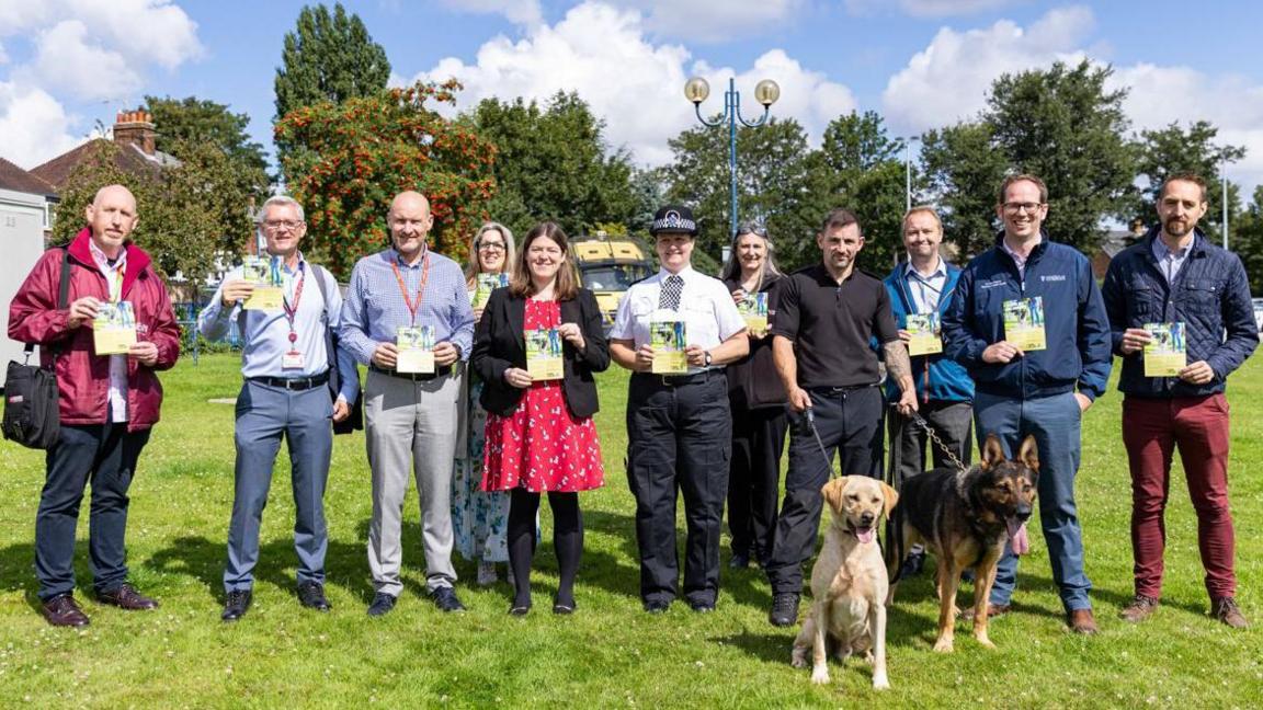 Taking The Lead campaign launch - Councillors and police represntatives and dog owners holding two dogs gather on grass and hold up campaign leaflets 