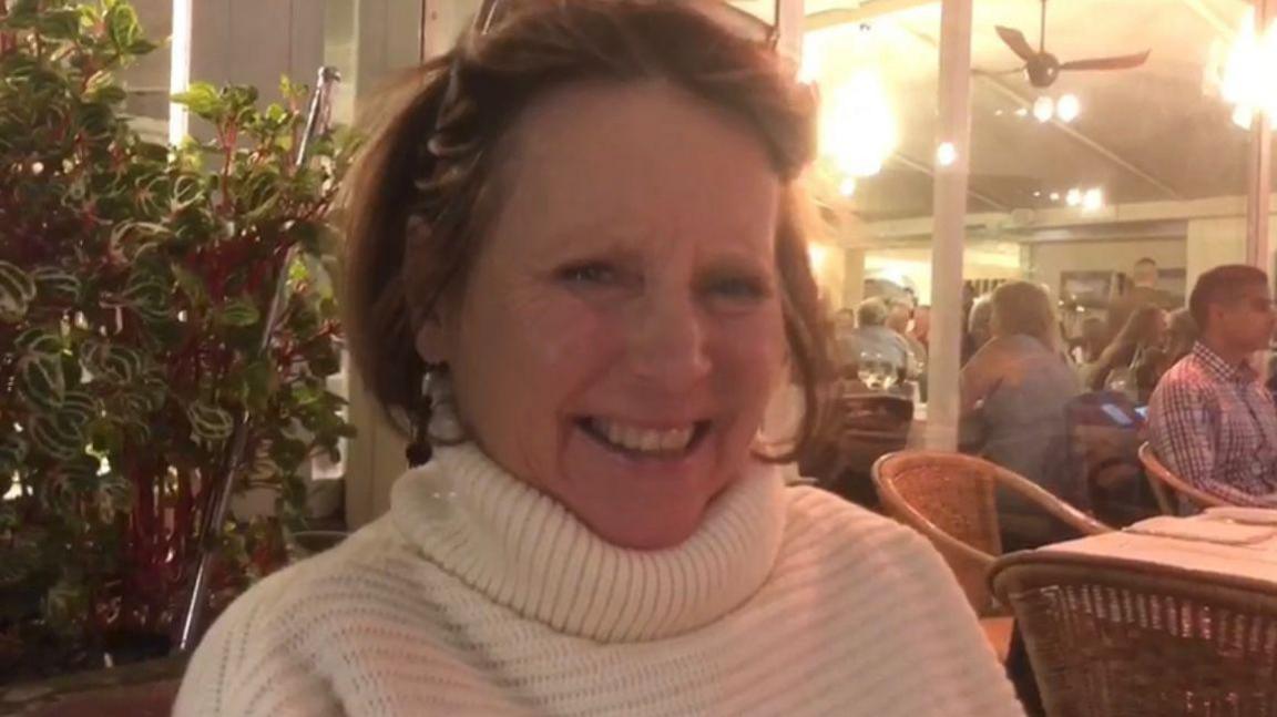 A woman uses her sunglasses to push her brown hair back as she smiles at the camera while sat in a restaurant with tables and people behind her.